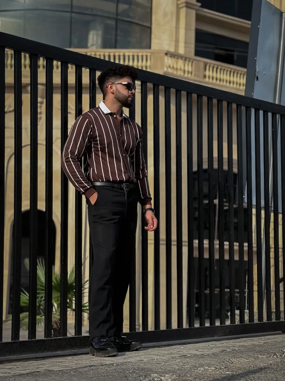 Man in a striped shirt and black pants standing in front of a black metal gate.
