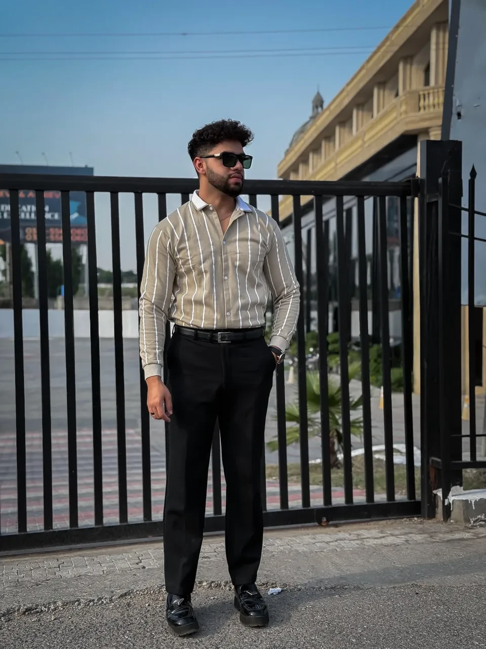 Man wearing sunglasses and a striped shirt standing in front of a black metal gate.