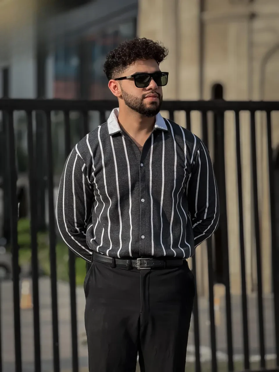 Man wearing a striped shirt and sunglasses standing in front of a black metal fence.