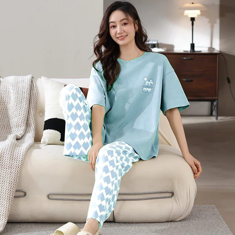 Woman wearing a light blue pajama set with a patterned design, sitting on a beige ottoman in a living room.