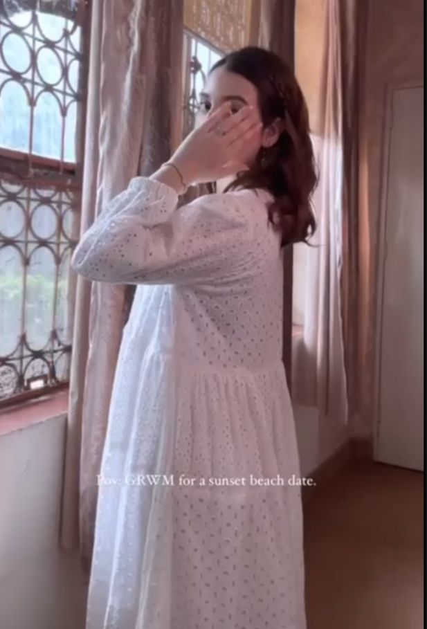 Woman in a white dress standing in a room with decorative curtains.