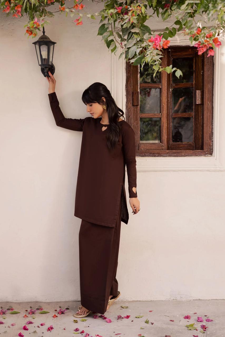 Woman in a brown outfit standing next to a white wall with flowers and a window in the background.