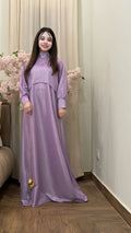 Woman wearing a long purple dress standing indoors with decorative flowers and a couch in the background.