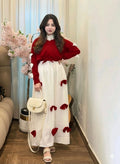 Woman in a red sweater and white dress with red accents standing in a room with flowers and furniture.