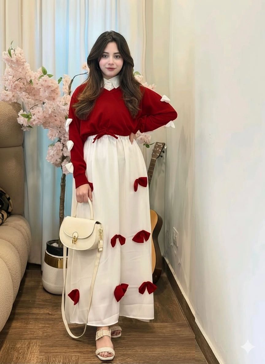 Woman in a red sweater and white dress with red accents standing in a room with flowers and furniture.