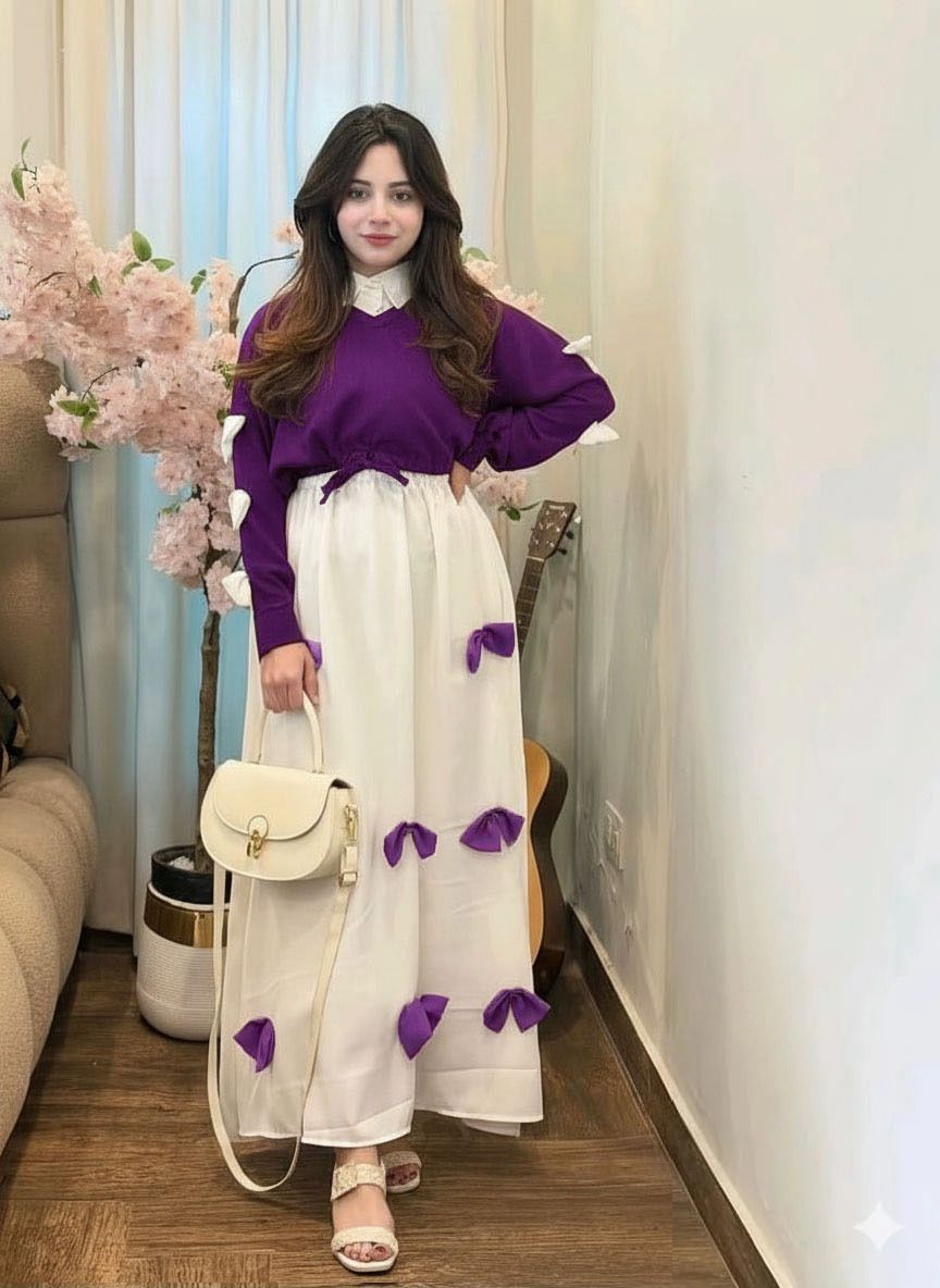 Woman in a purple sweater and white skirt with purple accents standing in a room with flowers and furniture.