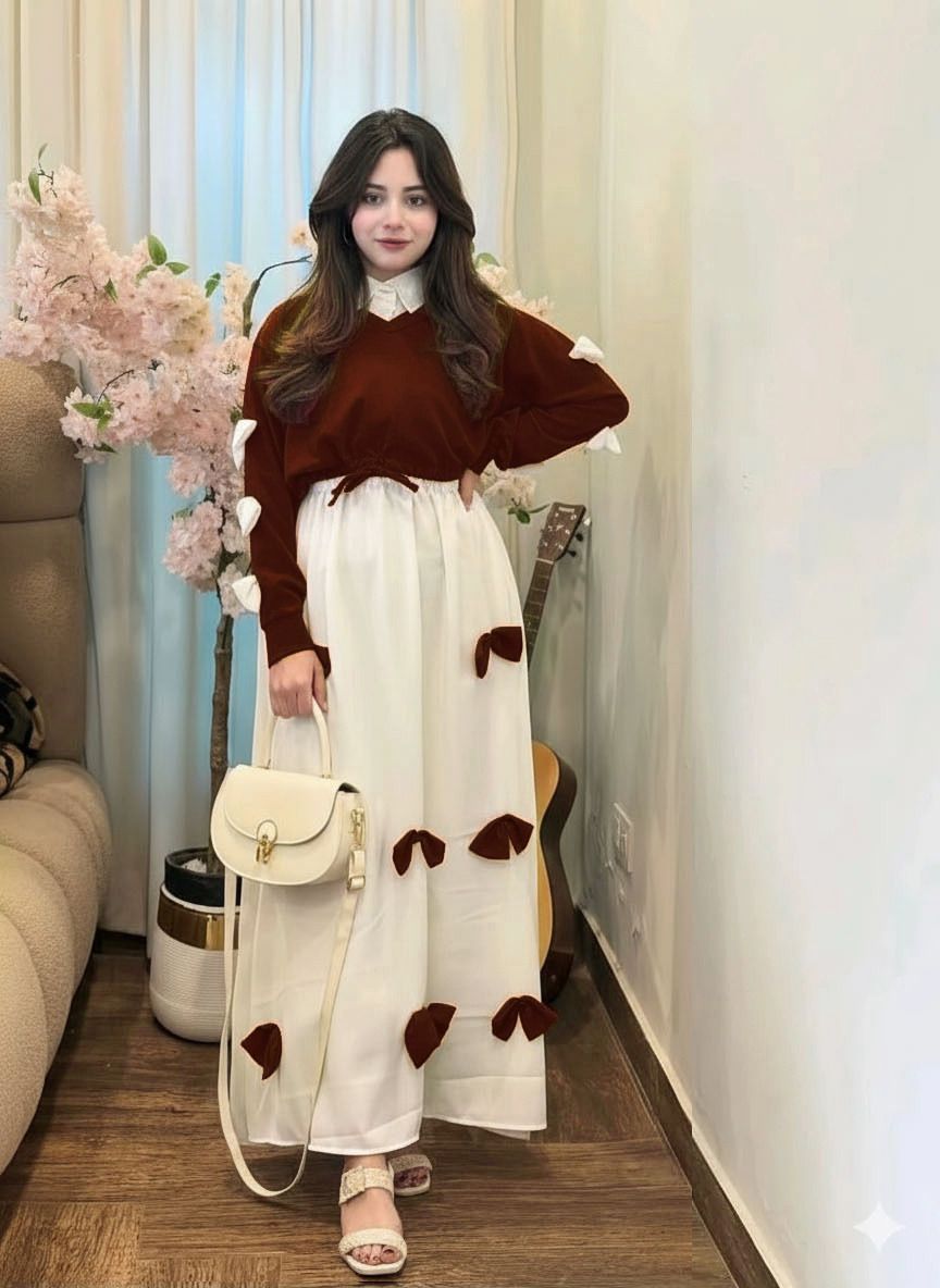 Woman in a brown sweater and white dress with brown polka dots standing in a room with flowers and furniture.