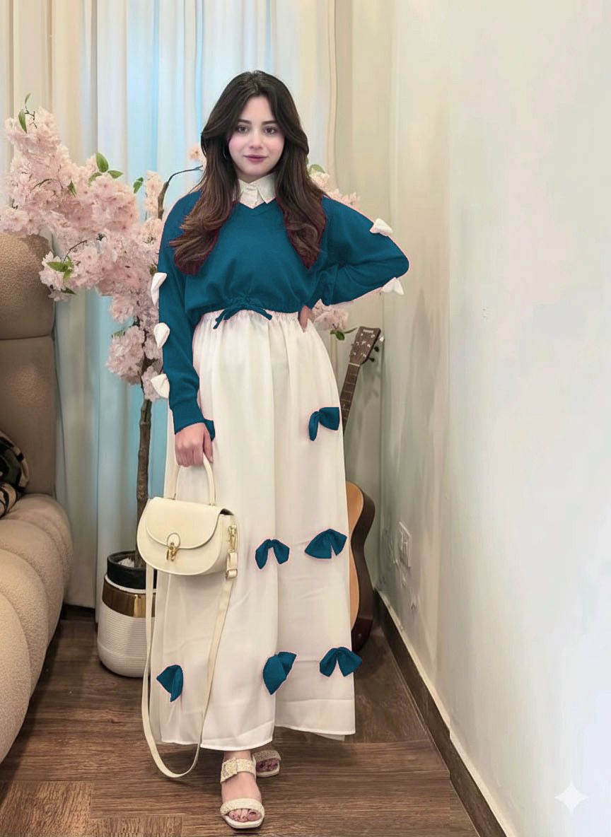 Woman in a teal sweater and white dress with blue floral patterns standing in a room with light-colored walls and a couch.