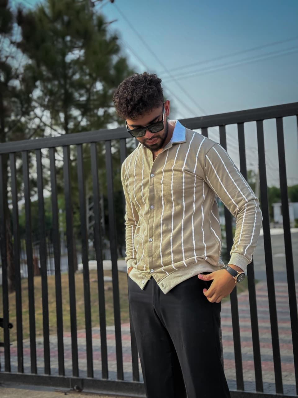 Man wearing a light-colored striped shirt and dark pants standing near a black metal fence.
