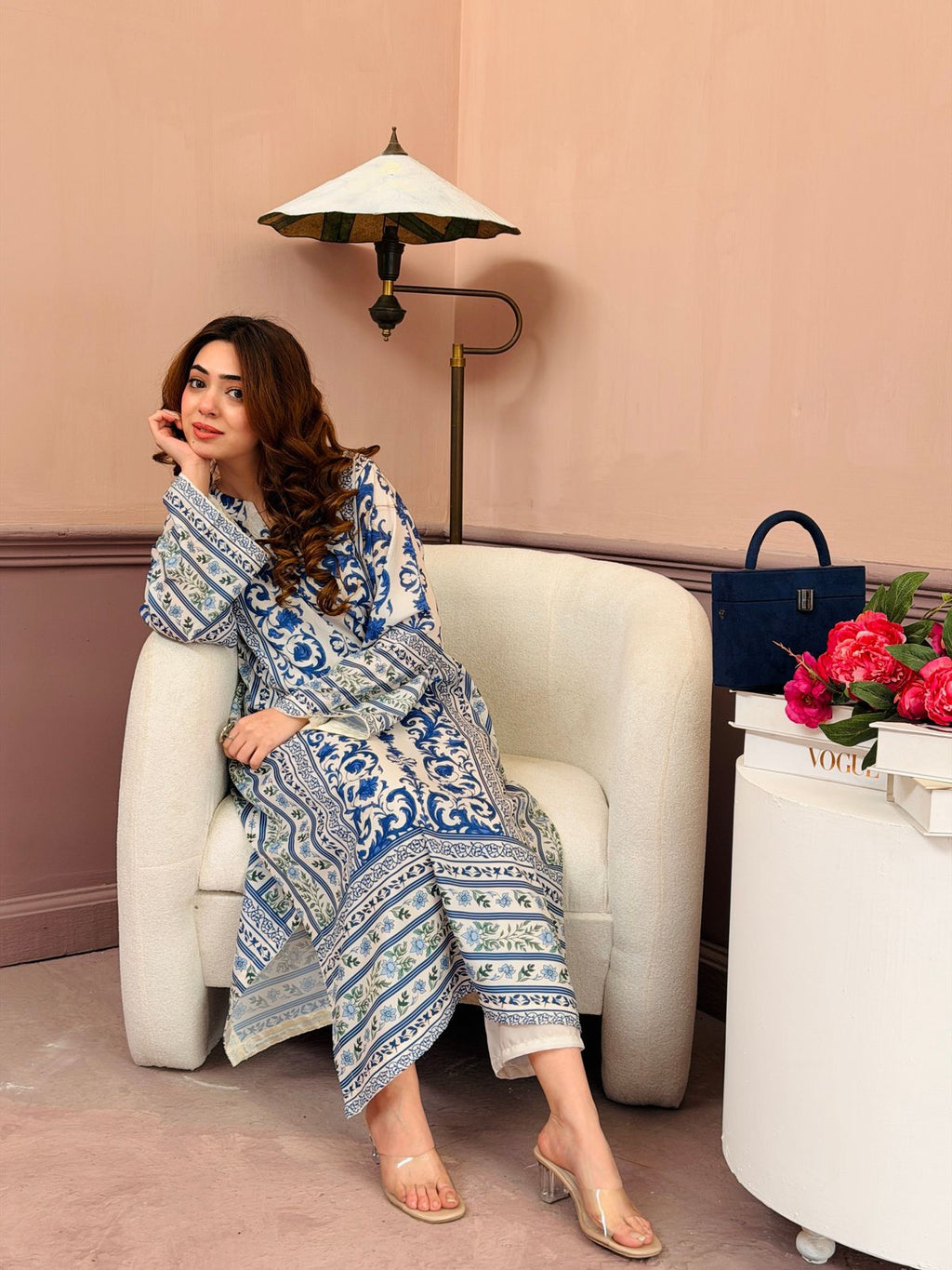 Woman in a blue and white patterned outfit sitting on a white chair with a pink wall and lamp in the background.