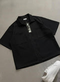 Black short-sleeve shirt with a tag on a gray surface, next to a white sneaker.