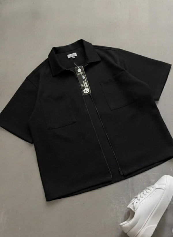 Black short-sleeve shirt with a tag on a gray surface, next to a white sneaker.