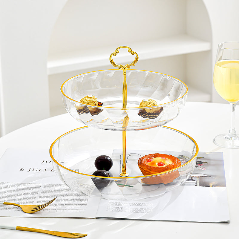Two-tiered glass serving tray with gold rim and handle, filled with small desserts on a white surface.