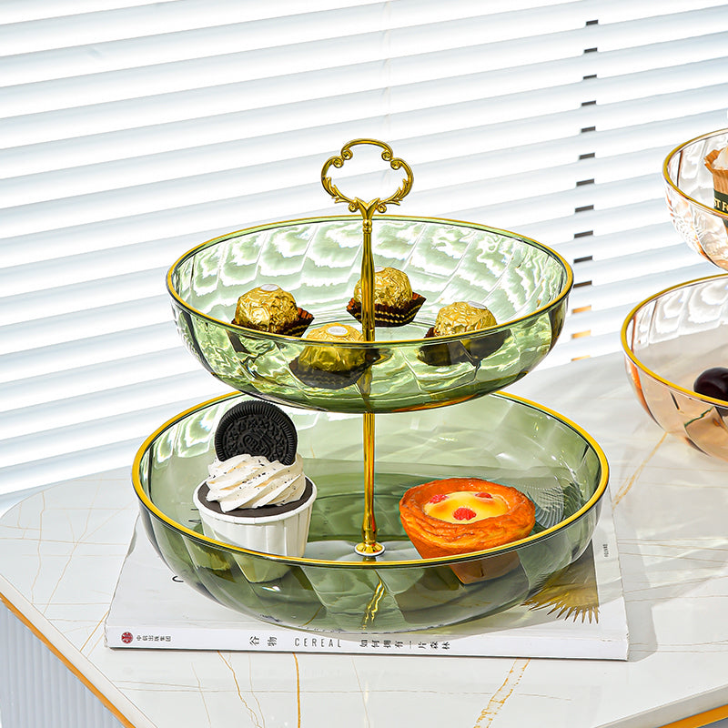 Two-tiered glass serving tray with gold accents displaying various desserts on a white surface.