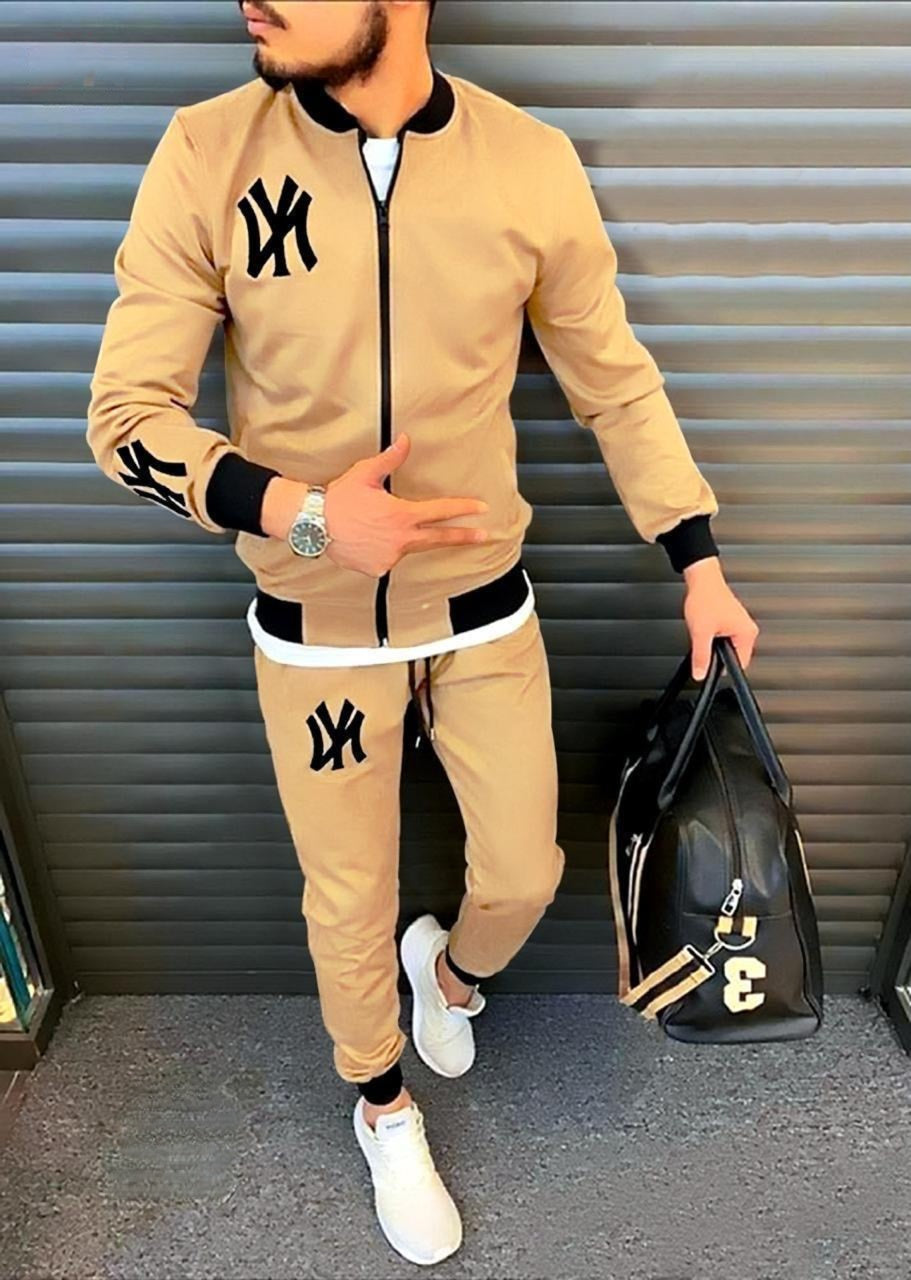 Man wearing a beige tracksuit with black accents, holding a black bag with gold lettering, against a metallic background.