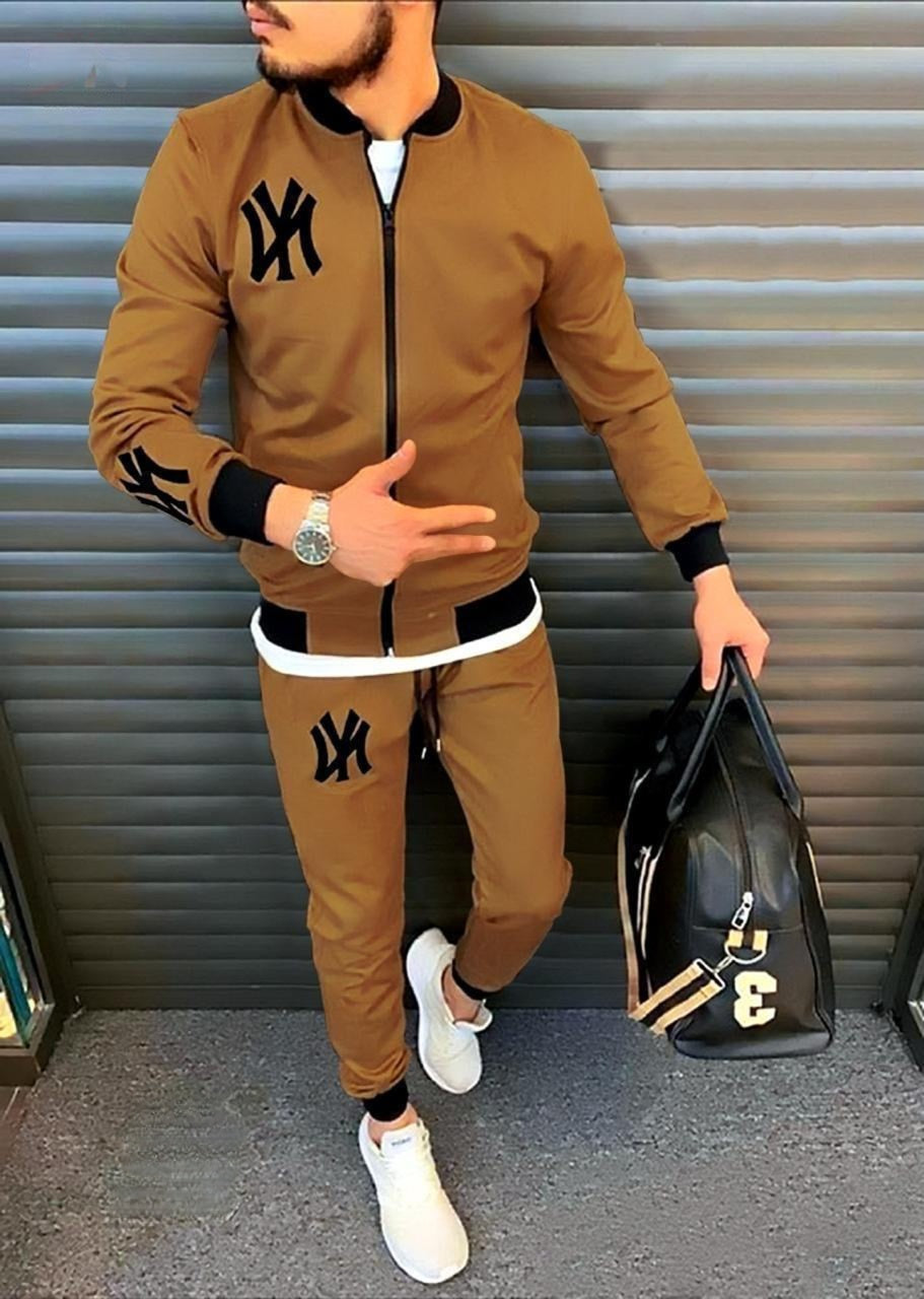 Man wearing a brown tracksuit with black logo, holding a black bag against a metallic background.
