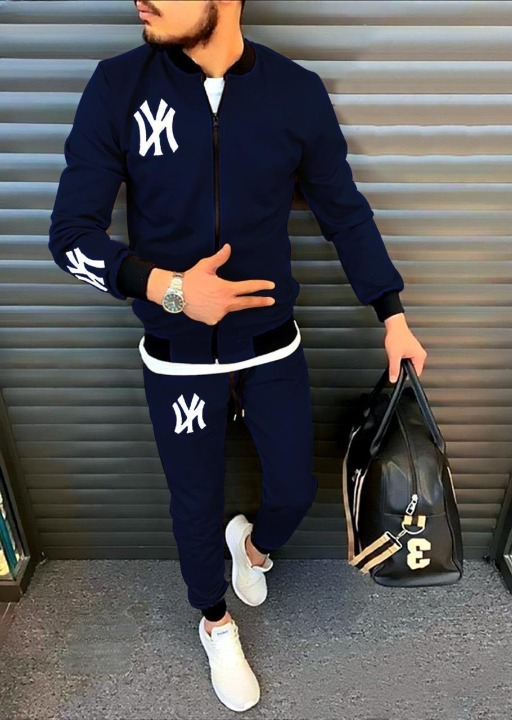 Person wearing a navy tracksuit with white logos, holding a black bag, standing against a metallic shutter.